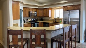 Une cuisine avec des meubles de rangement en bois, un réfrigérateur en acier inoxydable, un four à micro-ondes, une cuisinière et un évier. Il y a trois tabourets de bar en bois.
