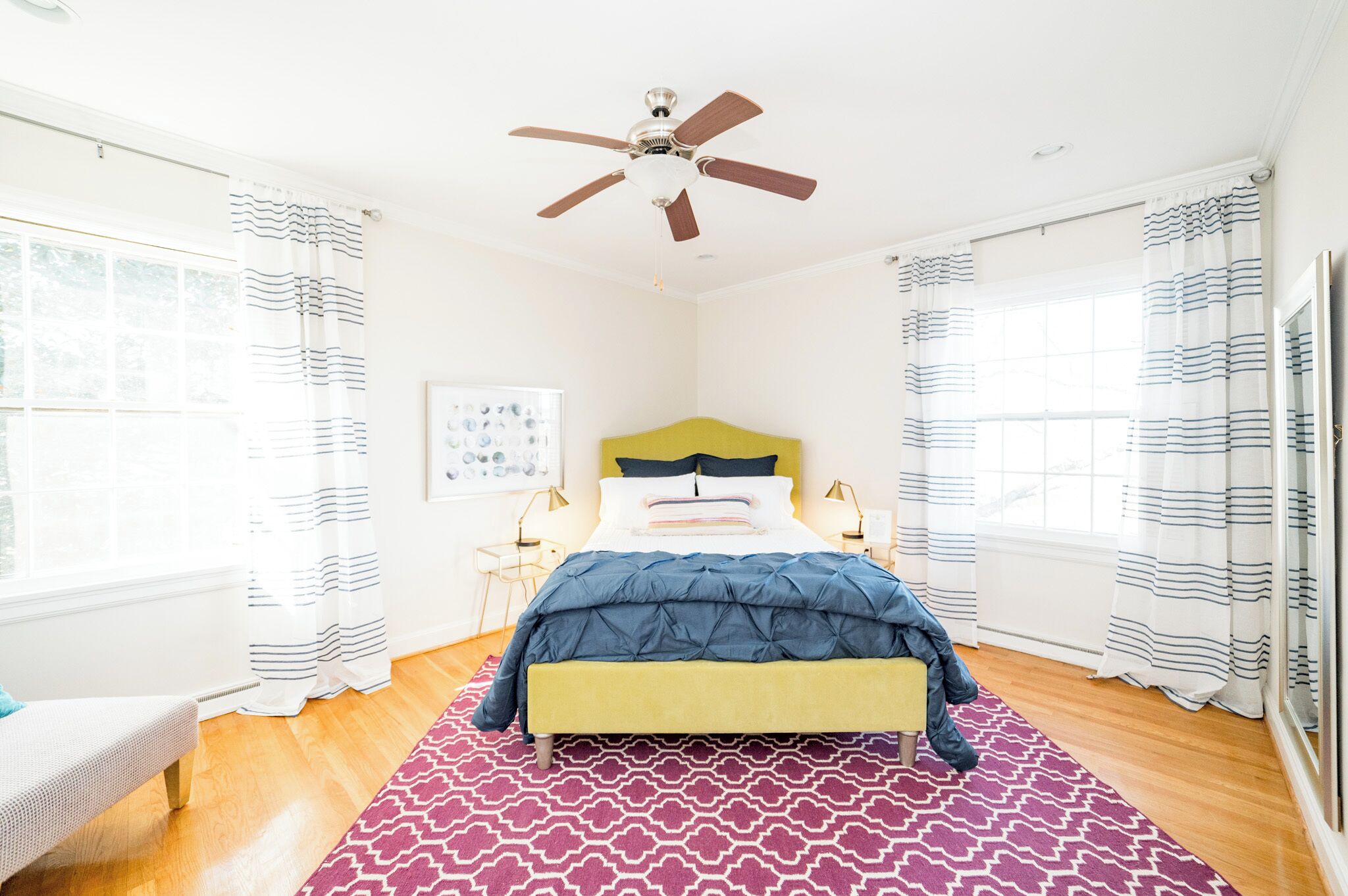 A bedroom with a bed, a ceiling fan, a window with curtains, a bedside table with a lamp, and a patterned rug.