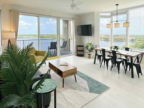 Un salón espacioso con un ventanal grande, una zona de comedor con mesa y sillas, y un balcón con vistas al agua.