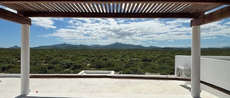 A rooftop terrace with a wooden pergola, white pillars, and a view of a mountainous landscape.