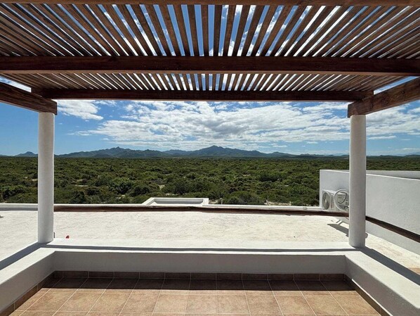 A rooftop terrace with a wooden pergola, white pillars, and a view of a mountainous landscape.