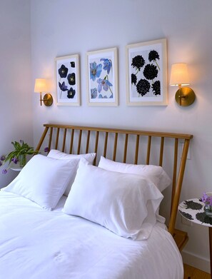 A neatly made bed with white linens, a wooden headboard, and three framed floral artworks on the wall.