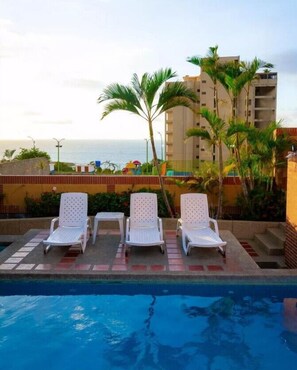 A poolside area with three white lounge chairs, a tiled deck, and a view of the ocean and a tall building in the background. - HOTEL MIRAMAR SUITES (Urimare)