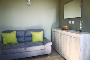 Un canapé gris avec deux coussins verts, un meuble en bois, un miroir et un diffuseur avec des bâtonnets en roseau.