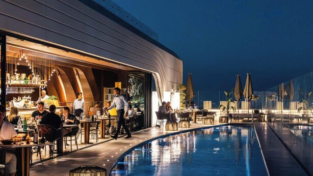 Ein Poolbereich mit Bar und Essbereich, ein Schwimmbecken und eine moderne Gebäudefassade.