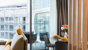 Ein modernes Hotelzimmer mit einem großen Panoramafenster, einer Couch mit Kissen, einem Esstisch mit Stühlen und einer Vase mit Blumen.