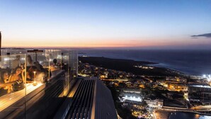 Un hôtel de grande hauteur offrant une vue panoramique sur la côte, un bar sur le toit doté de rampes en verre, et une vue sur la ville en arrière-plan.