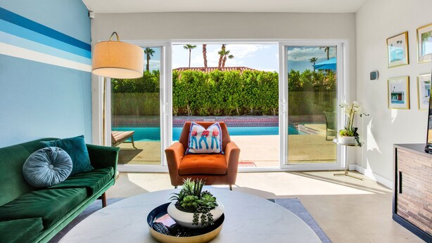 A modern living room with a green sofa, a round coffee table with a plant, a chair with a colorful cushion, and a view of a swimming pool through large glass doors. - Stunningly designed, colorful, fun 3-bedroom house in Central Palm Springs. (Palm Springs)
