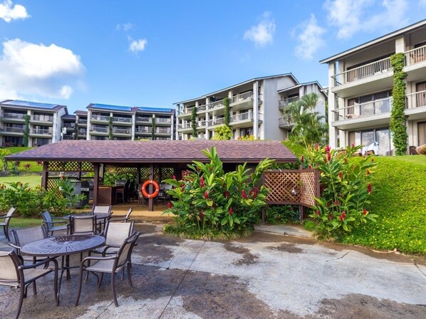 A covered outdoor seating area with chairs and a table, surrounded by lush greenery and a clear sky. - Ocean View Studio at HBR, A/C Included (Princeville)