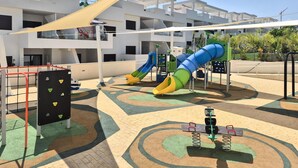 A playground with slides and climbing structures, surrounded by a patterned pavement and a building with balconies in the background. - Cozy apartment in Finestrat with WiFi (Finestrat)