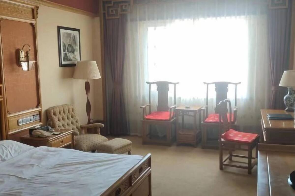 Een hotelkamer met een bed, twee fauteuils, een tafeltje, een lamp, een schilderij aan de muur en een raam met gordijnen.