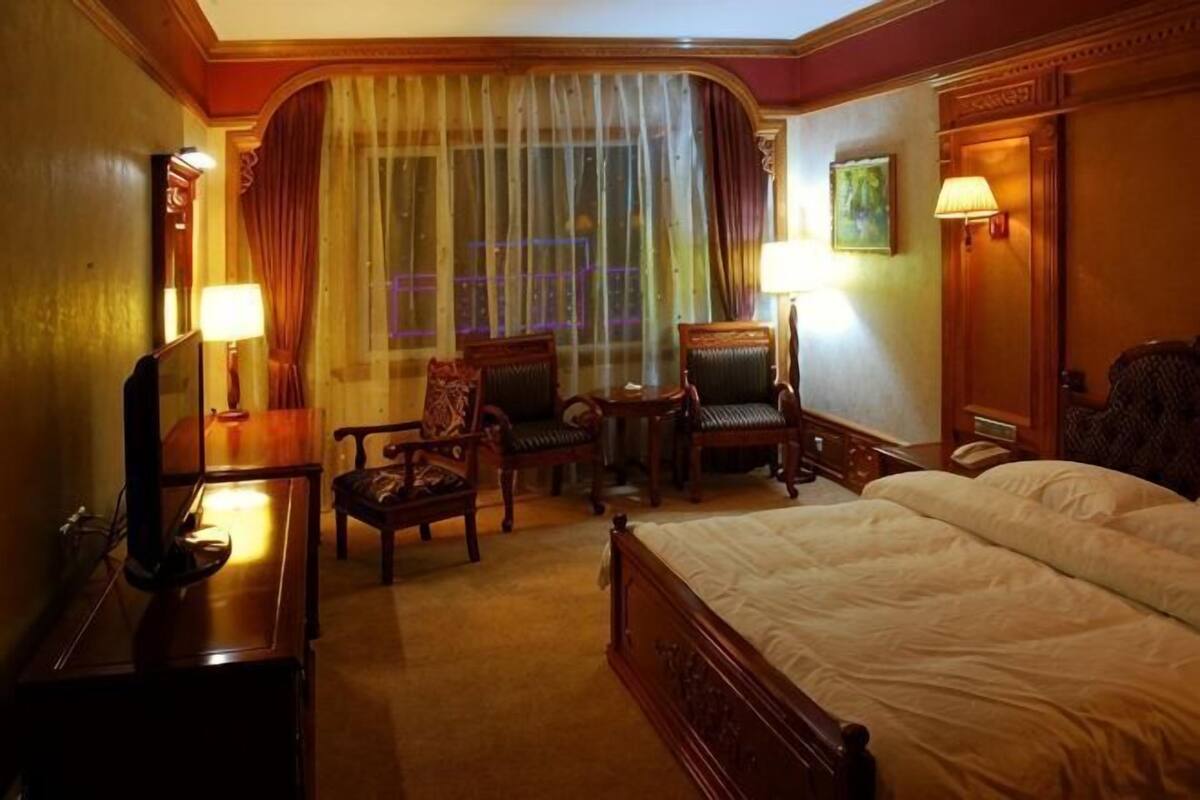 Hotelkamer met een bed, twee stoelen, een klein tafeltje, een spiegel, een lamp en een raam met gordijnen.