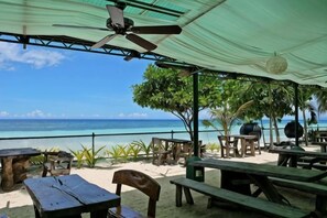 Eine Terrasse direkt am Strand mit Holztischen und -stühlen, einem Deckenventilator und Blick auf den Ozean.