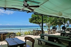 Une terrasse au bord de la plage, avec des tables et des chaises en bois, un ventilateur de plafond et une vue sur l’océan.