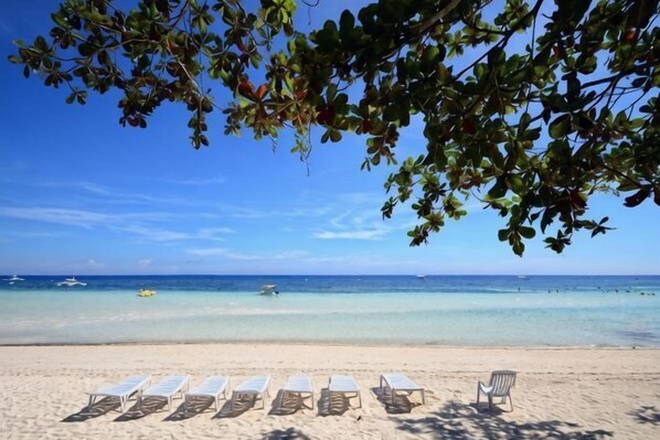 Une plage avec des chaises longues blanches et une seule chaise placée sous un arbre, d’où l’on a une vue sur la mer.