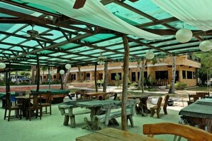 Un espace extérieur aménagé pour les repas, avec des tables et des bancs en bois, une structure couverte dotée d’un toit, et un bâtiment visible à l’arrière-plan.