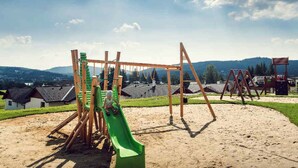 A playground with a slide and swings, set against a backdrop of houses and mountains.