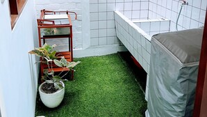 A small, enclosed bathroom with a white tiled wall, a green artificial grass floor, a wooden shelf with plants, and a bathtub covered with a grey cloth.