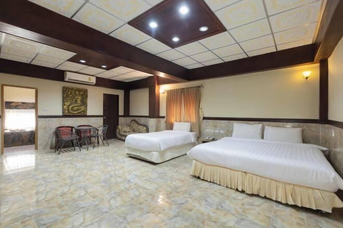 Quarto de hotel com duas camas, área de refeições com cadeiras, teto com iluminação embutida e ar-condicionado fixado na parede.