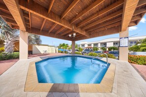 Un espace piscine agrémenté d’une pergola en bois, d’une terrasse carrelée et offrant une vue sur un bâtiment situé au loin.