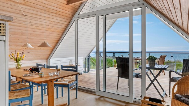 Un intérieur moderne avec un plafond en bois, un coin repas avec des chaises bleues et une grande porte vitrée donnant sur une terrasse extérieure avec vue sur la mer.