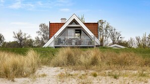 Une maison moderne avec un toit triangulaire, un balcon avec des chaises et un espace sablonneux devant.