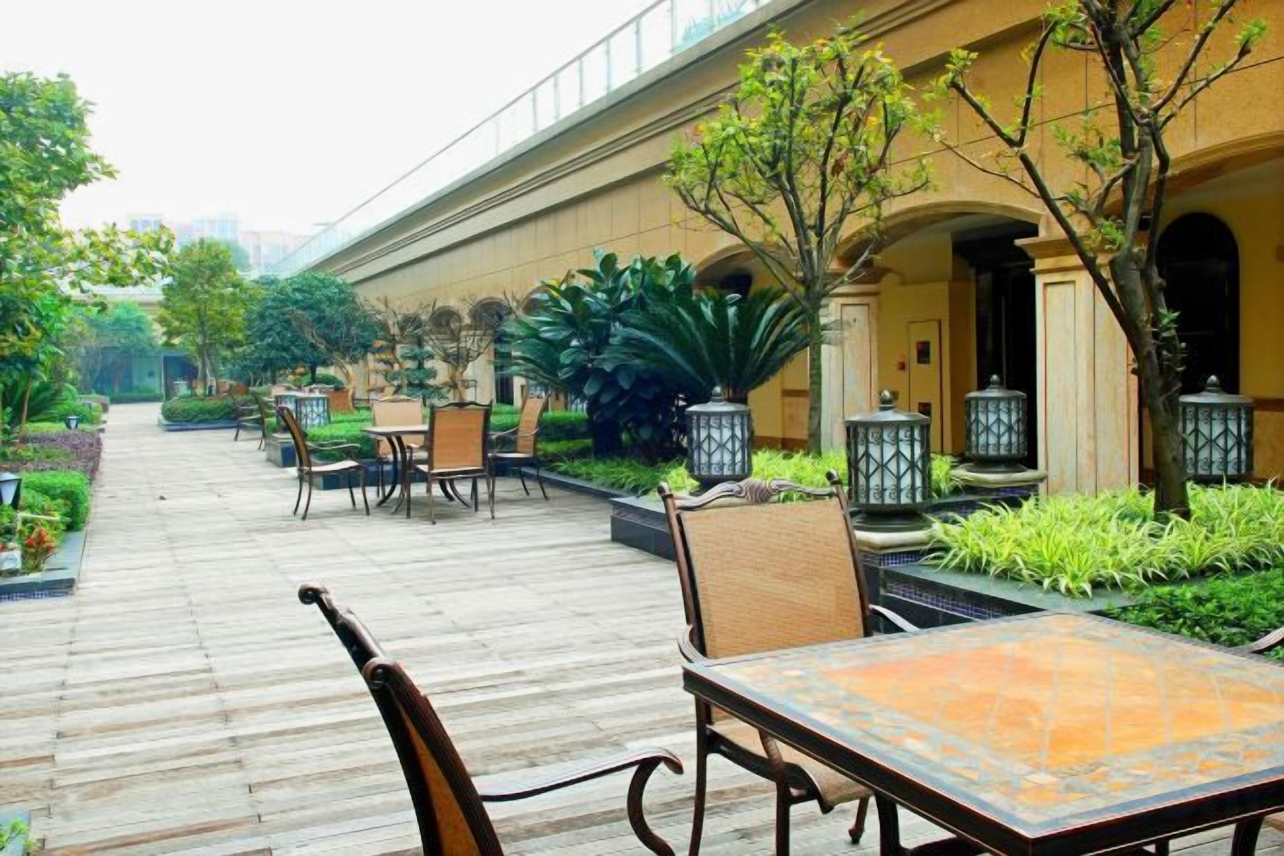 An outdoor seating area with tables and chairs, surrounded by greenery and architectural columns.