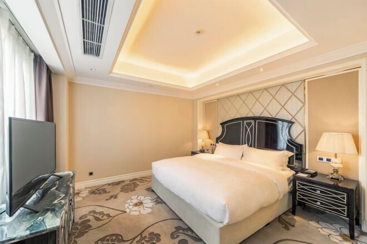 A hotel room with a large bed, a desk with a flat-screen TV, a nightstand with a lamp, and a decorative mirror.