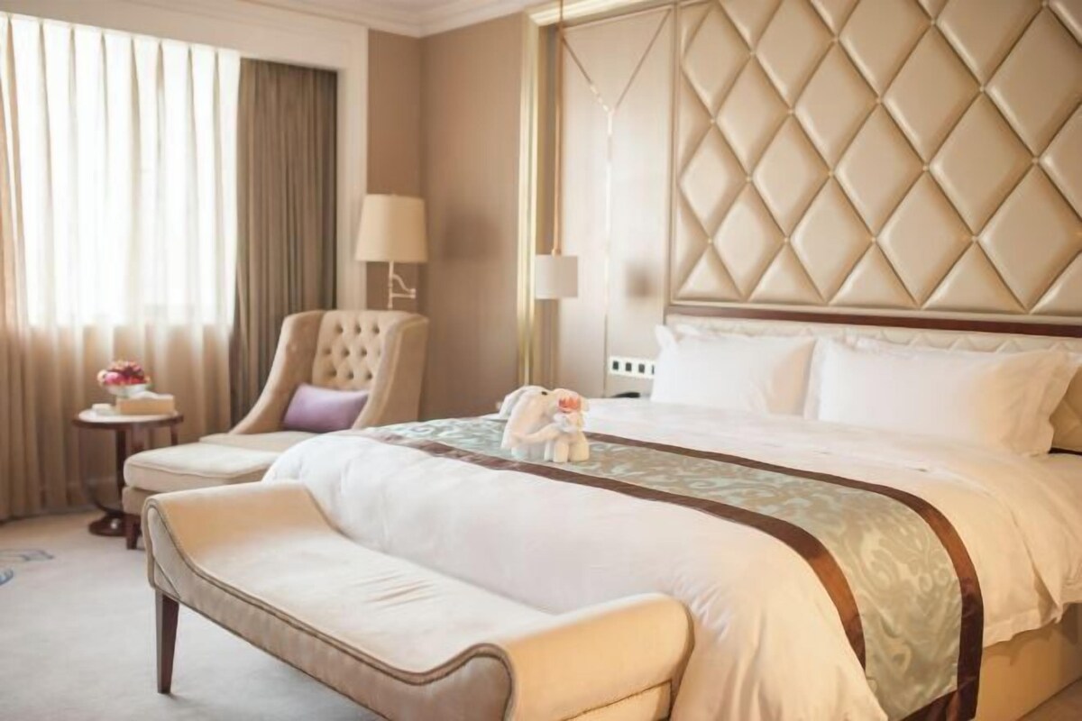 A hotel room with a large bed, a tufted headboard, a bench, a chair, and a nightstand with a lamp.