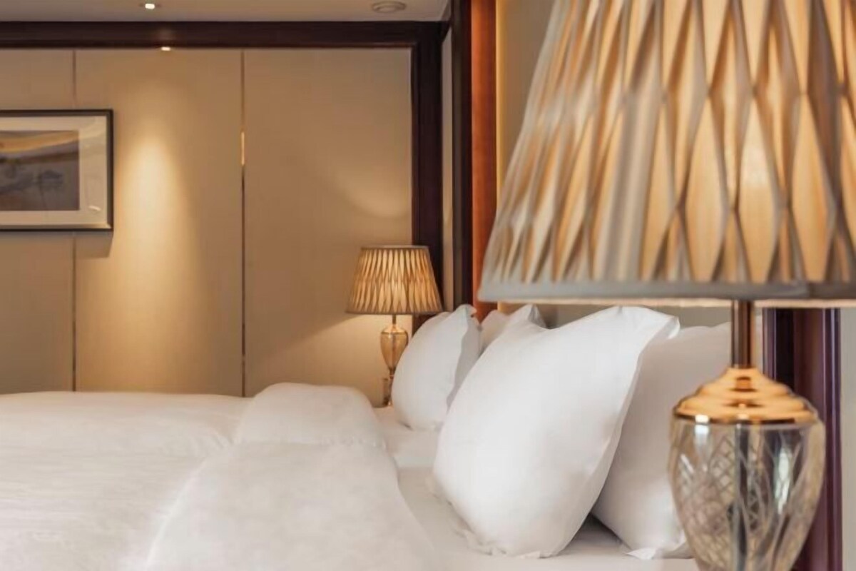 A neatly made bed with white linens and pillows, a wall-mounted lamp, and a framed picture on the wall.