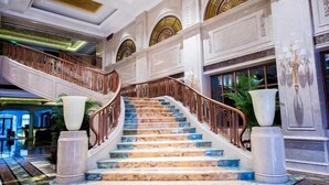 Un magnifique escalier en marbre, agrémenté d’une rampe en bois, mène à un hall d’entrée orné de grandes plantes en pot et doté d’un plafond haut orné de motifs complexes.