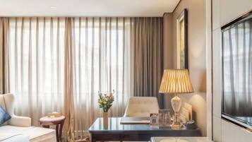 A hotel room with a bed, a desk with a lamp, a chair, a small table with a vase of flowers, and a large window with curtains.