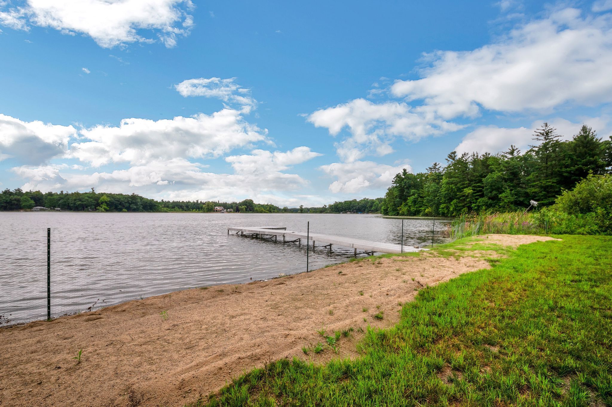 A lakeside view with a sandy beach, a wooden dock, and a forested area in the background.