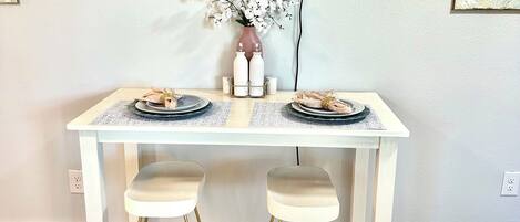 A white dining table with two stools, a vase with white flowers, and a flat-screen TV mounted on the wall.