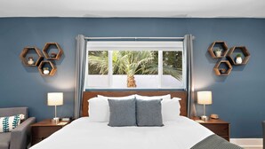 A hotel room with a large bed, two bedside tables, a nightstand, a chair, and a window with a view of palm trees. - Modern Palm Springs Pool Home Near Downtown Sleeps 8 (Palm Springs)