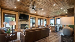 A spacious living area with a fireplace, wooden ceiling, and large windows. - Broken Bow Cabin w/ Mountain Views & Sunset Vibes (Broken Bow)