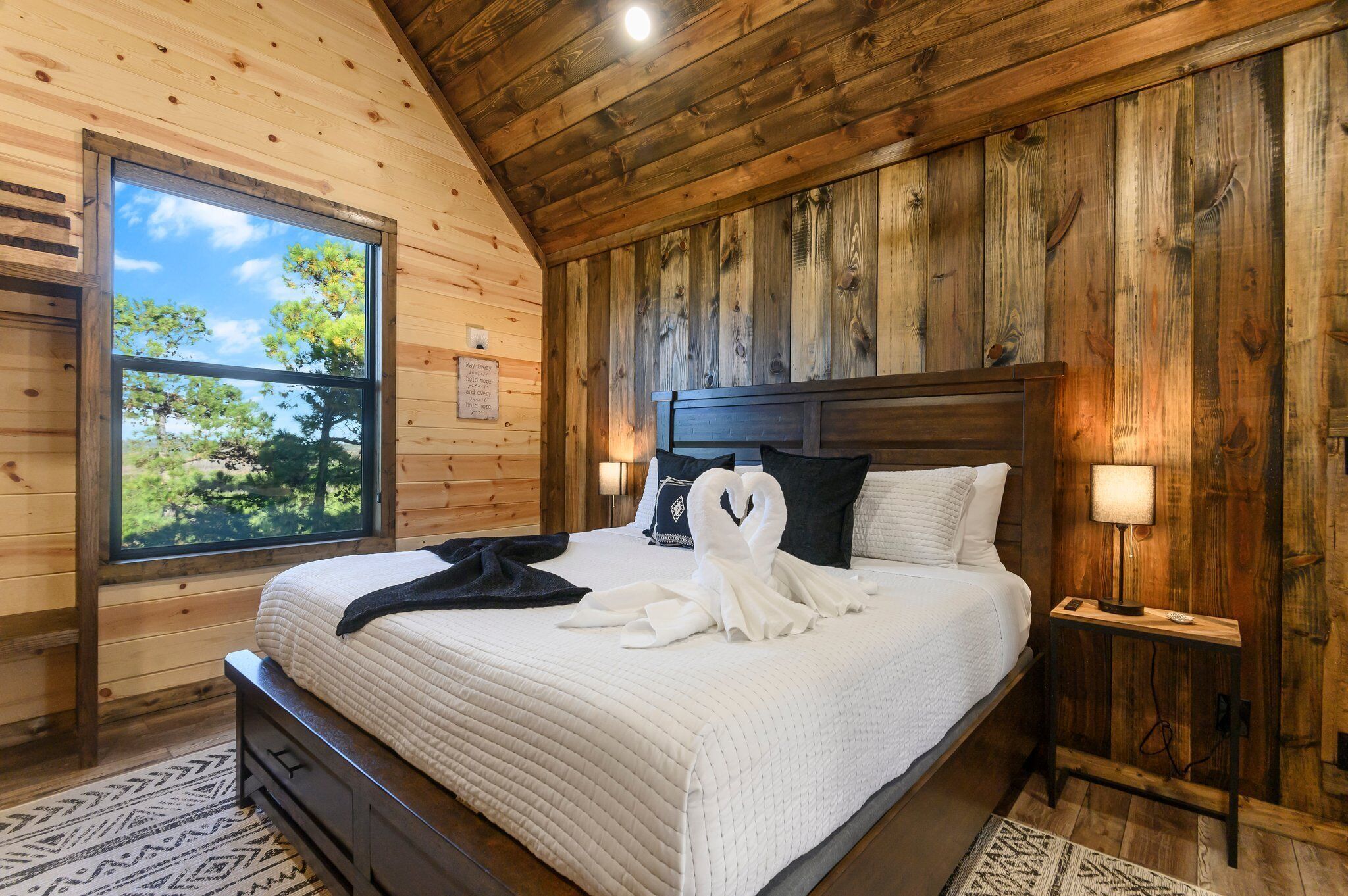 A wooden-paneled bedroom with a large window, a bed with white bedding, two bedside lamps, and a wooden nightstand.