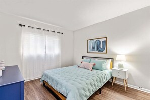 Une chambre à coucher avec un lit, une table de chevet avec une lampe, une œuvre d’art accrochée au mur et une fenêtre avec des rideaux légers.