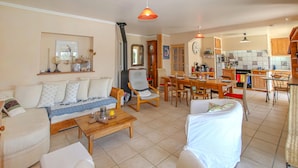Ein geräumiges Wohnzimmer mit Sofa, Sesseln, einem Couchtisch und einem Essbereich mit Tisch und Stühlen. Im Hintergrund ist eine Küche erkennbar.