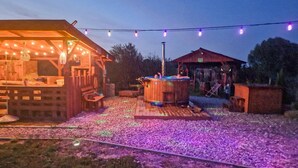 Eine Holzhütte mit einer Terrasse und einem Whirlpool, umgeben von einem Kiesbereich mit violetter Beleuchtung.