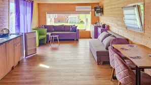 Ein gemütliches Blockhaus-Interieur mit Holzboden, einer Couch, einem Esstisch mit Stühlen, einer Küchenzeile und einem großen Fenster.
