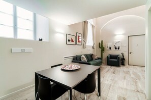 Ein modernes Wohnzimmer mit Esstisch, Sofa und Sessel. Ein Fenster mit Vorhängen, eine Tür und eine Säulencactus stehen auf dem Boden.