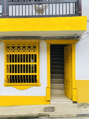 Un edificio con fachada amarilla, una ventana con barrotes y una escalera que lleva a un nivel superior.
