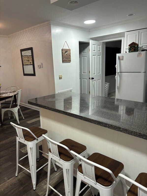 A kitchen with a granite countertop, white chairs, a refrigerator, and a door leading to another room.