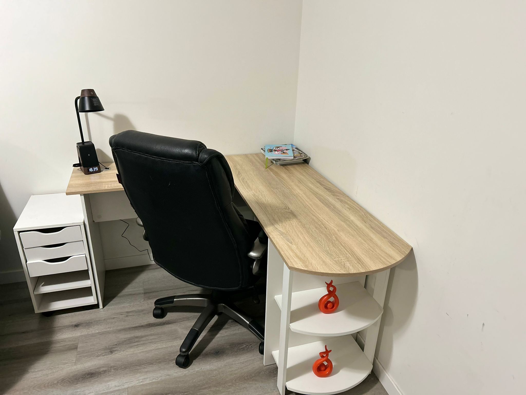 Um pequeno espaço de escritório com uma escrivaninha de madeira, uma cadeira de escritório preta, uma estante branca e uma prateleira com objetos decorativos vermelhos.