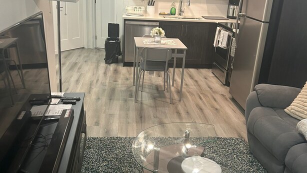 A modern living room with a flat-screen TV, a glass coffee table, and a grey sofa. The kitchen area is visible in the background with dark wooden cabinets and stainless steel appliances. - Cozy and Private Suite - Free On-Prem Parking & PS5 Available (Calgary)
