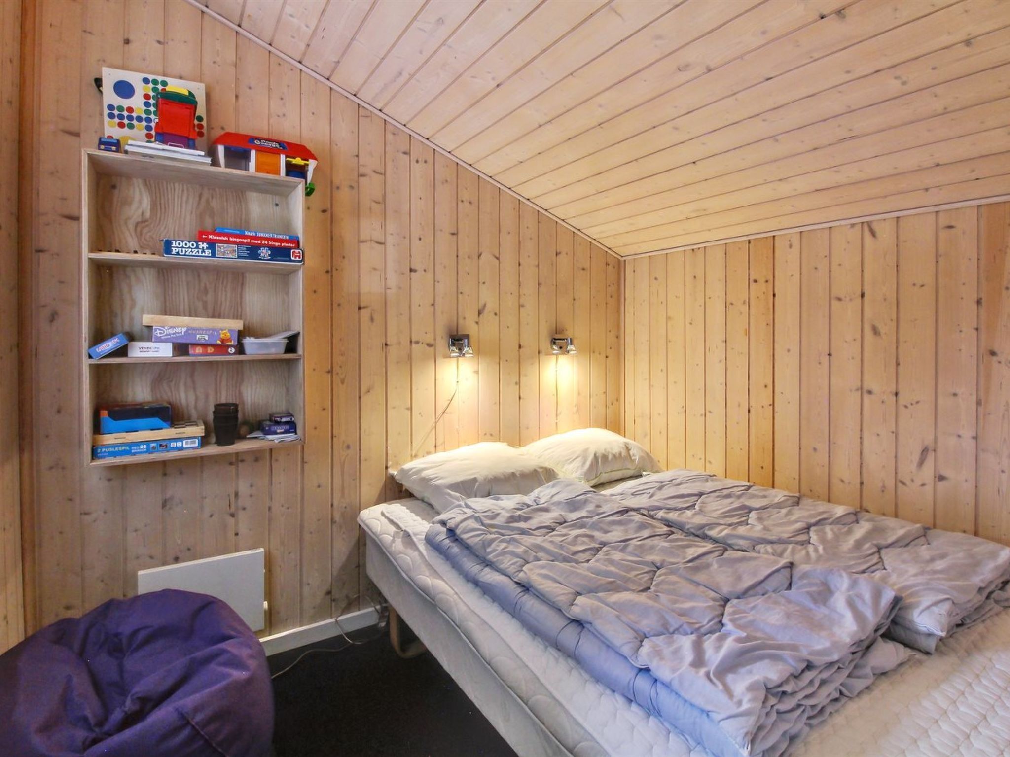 Ein kleiner Raum mit Holzvertäfelung, einem Einzelbett, einem Regal mit diversen Gegenständen und zwei Wandleuchten.