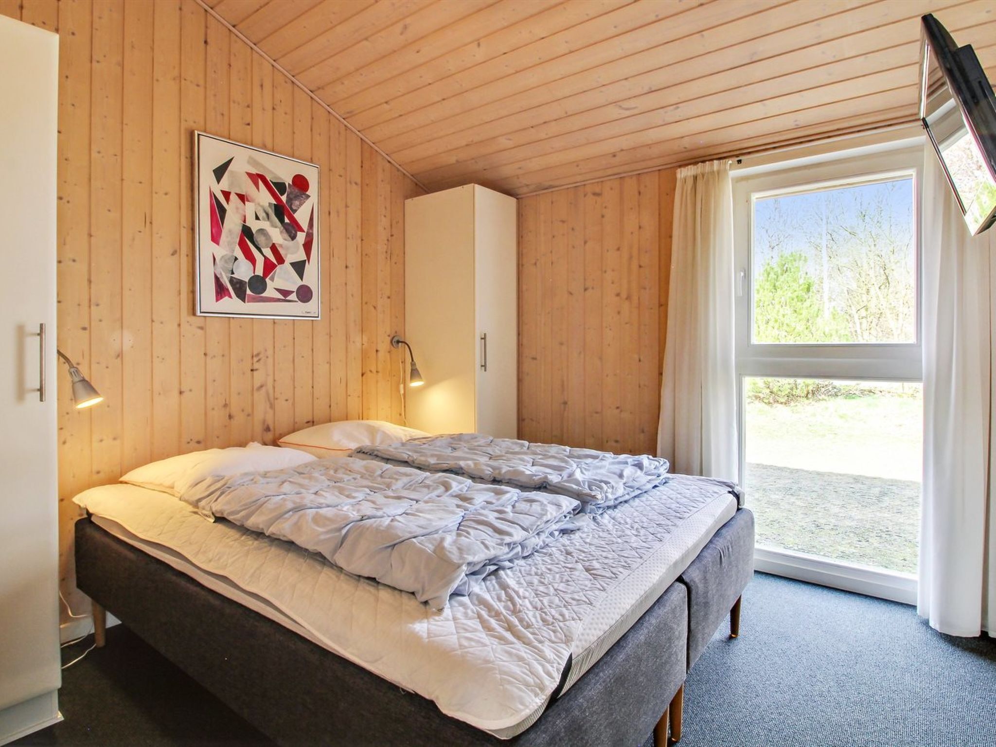 A bedroom with a bed, a wooden ceiling, a window, and a wall-mounted picture.