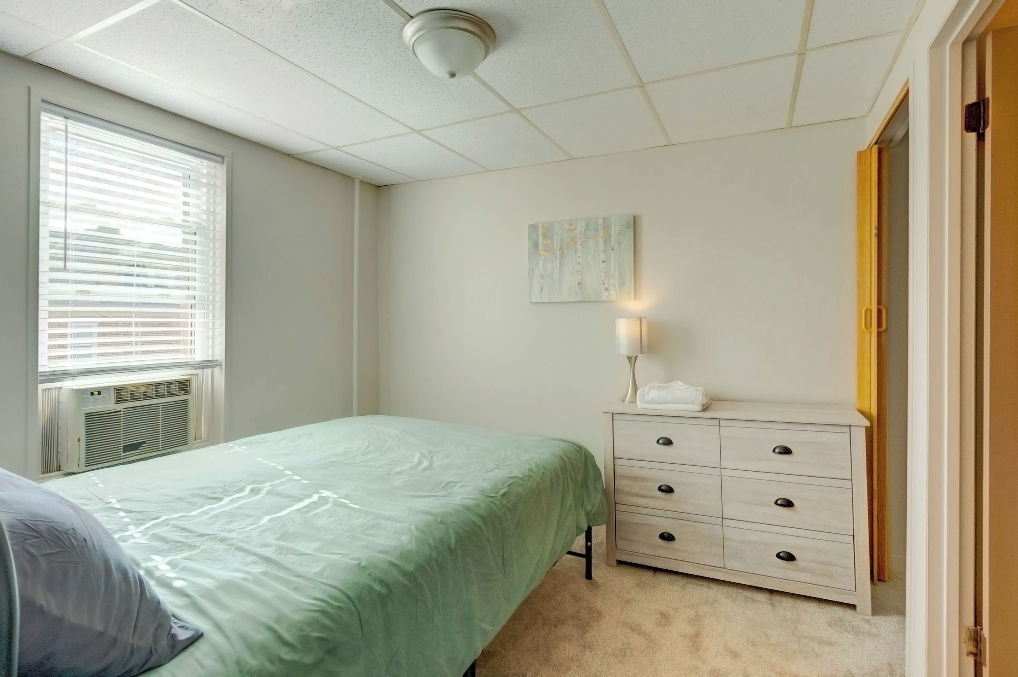 Une chambre à coucher comprenant un lit, une commode, une table de chevet, une fenêtre avec des stores et un luminaire au plafond.