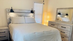 Una camera da letto con una testiera in legno, un letto con lenzuola bianche, un comodino, una cassettiera e uno specchio con una lampada da comodino.